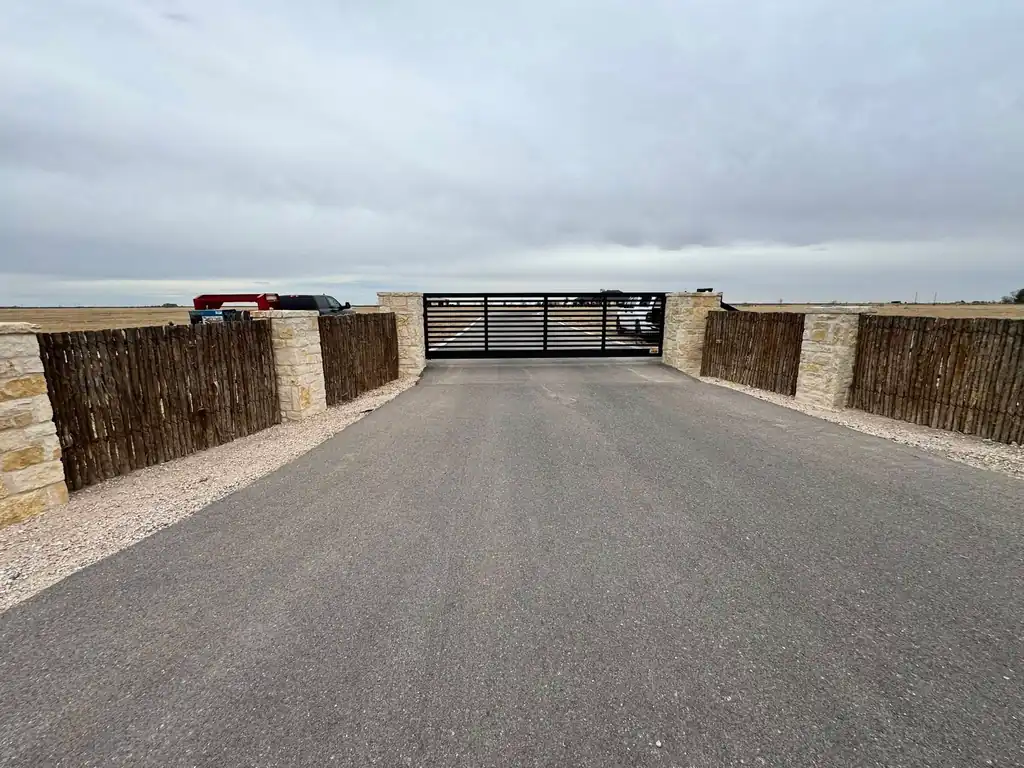 A slide gate approach between limestone columns with cedar-post fence wings, seen head on down a long drive