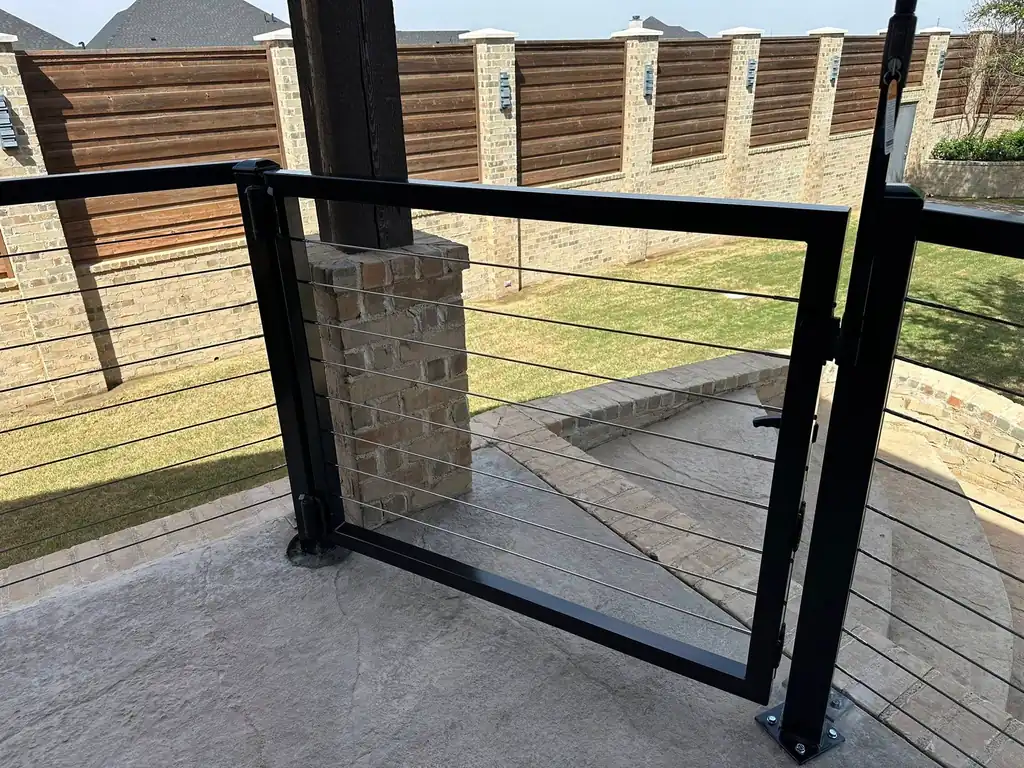 A black cable-rail garden gate against a run of horizontal cedar and brick fence